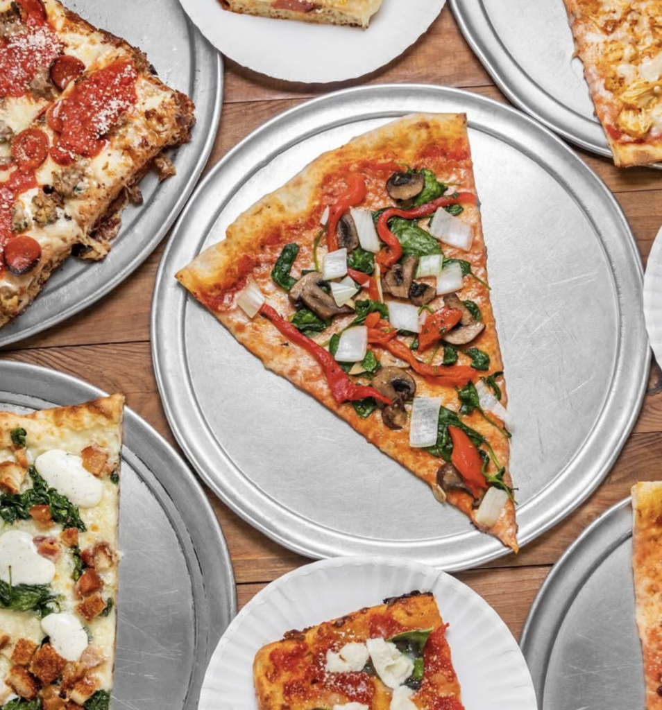 A variety of pizzas on round trays and paper plates, featuring different toppings like pepperoni, vegetables, and cheese, are arranged on a wooden surface.